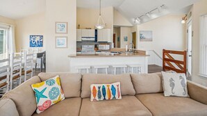 Un soggiorno moderno con un divano color beige, cuscini colorati, una cucina sullo sfondo e una sedia in legno.