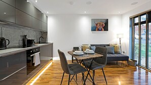 Una cocina moderna con armarios oscuros, un comedor con mesa y sillas de madera, y un sofá con vistas a través de grandes ventanales.