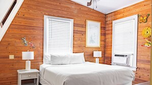 A bedroom with a wooden wall, a bed with white bedding, two bedside tables with lamps, and a window with blinds.