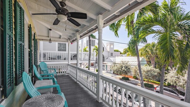A covered porch with wicker furniture, a ceiling fan, and a view of a street with palm trees and buildings.