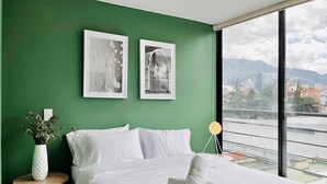 Une chambre d’hôtel moderne avec un mur vert, un grand lit recouvert de draps blancs, deux œuvres d’art encadrées, une table de chevet avec un vase, et une vue sur la ville à travers de grandes fenêtres.