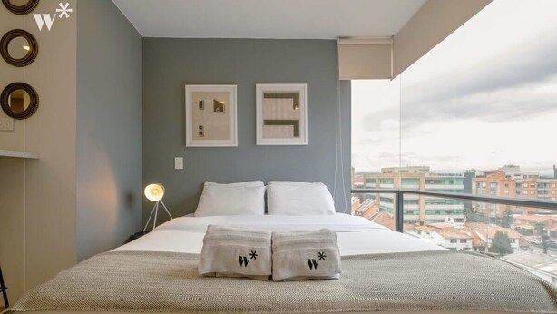 Hotelkamer met een bed, twee afbeeldingen in lijsten aan de muur, een balkon met uitzicht op de stad, en handdoeken met een logo op het bed.