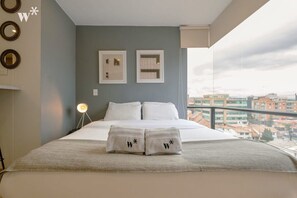 Habitación de hotel con cama, dos cuadros en la pared, un balcón con vista a la ciudad y toallas con un logotipo en la cama.