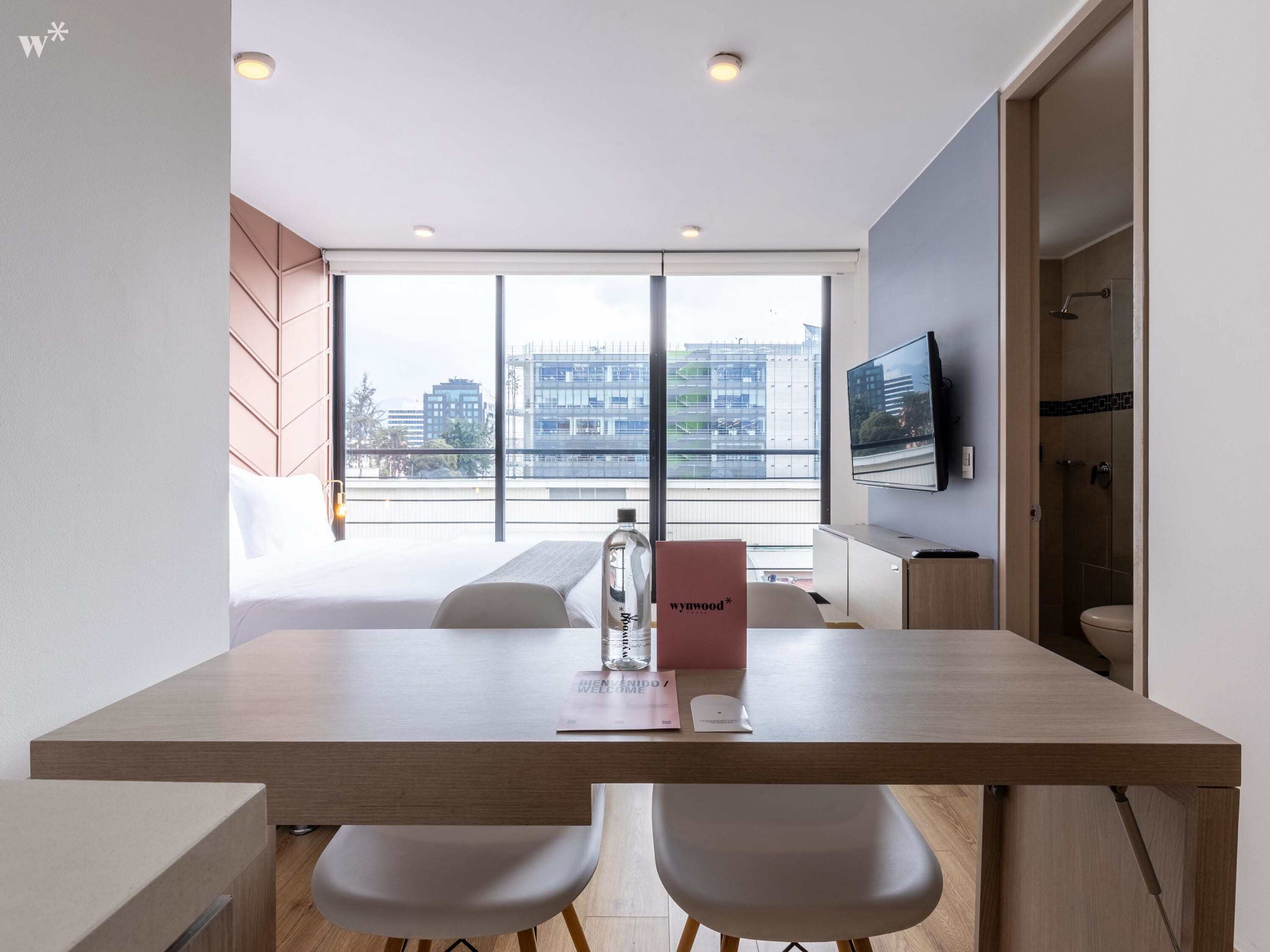 Ett modernt hotellrum med ett matbord i trä, två vita stolar, en platt-TV som är monterad på väggen och ett stort fönster med utsikt över staden.