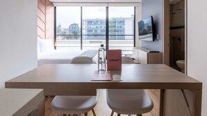 Une chambre d’hôtel moderne avec une table à manger en bois, deux chaises blanches, un téléviseur à écran plat fixé au mur et une grande fenêtre donnant sur la ville.