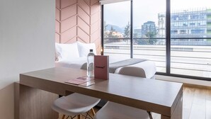 Une chambre d’hôtel moderne dotée d’une grande fenêtre donnant sur la ville, d’un bureau en bois avec une chaise et d’un lit avec une literie blanche.