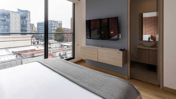 Une chambre à coucher moderne, dotée d’un grand lit, d’un téléviseur fixé au mur et offrant une vue sur le paysage urbain grâce à de grandes fenêtres.