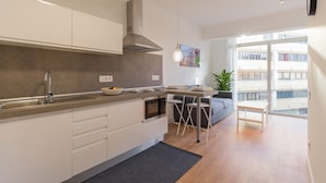 Eine moderne Küche mit einer zentralen Kochinsel, ein Essbereich mit grauem Sofa und Blick auf ein Gebäude durch große Fenster.