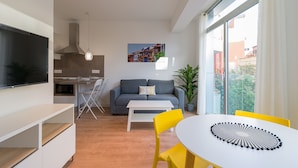 Ein modernes Wohnzimmer mit einer Couch, einem Esstisch mit gelben Stühlen, einem an der Wand montierten Fernseher und einer Küche im Hintergrund.