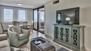 A living room with a sofa, ottoman, and a TV on a cabinet. There is a dining area with chairs and a table. - Vacation and relax in a recently renovated two bedroom condo. One free beach cha (Destin)