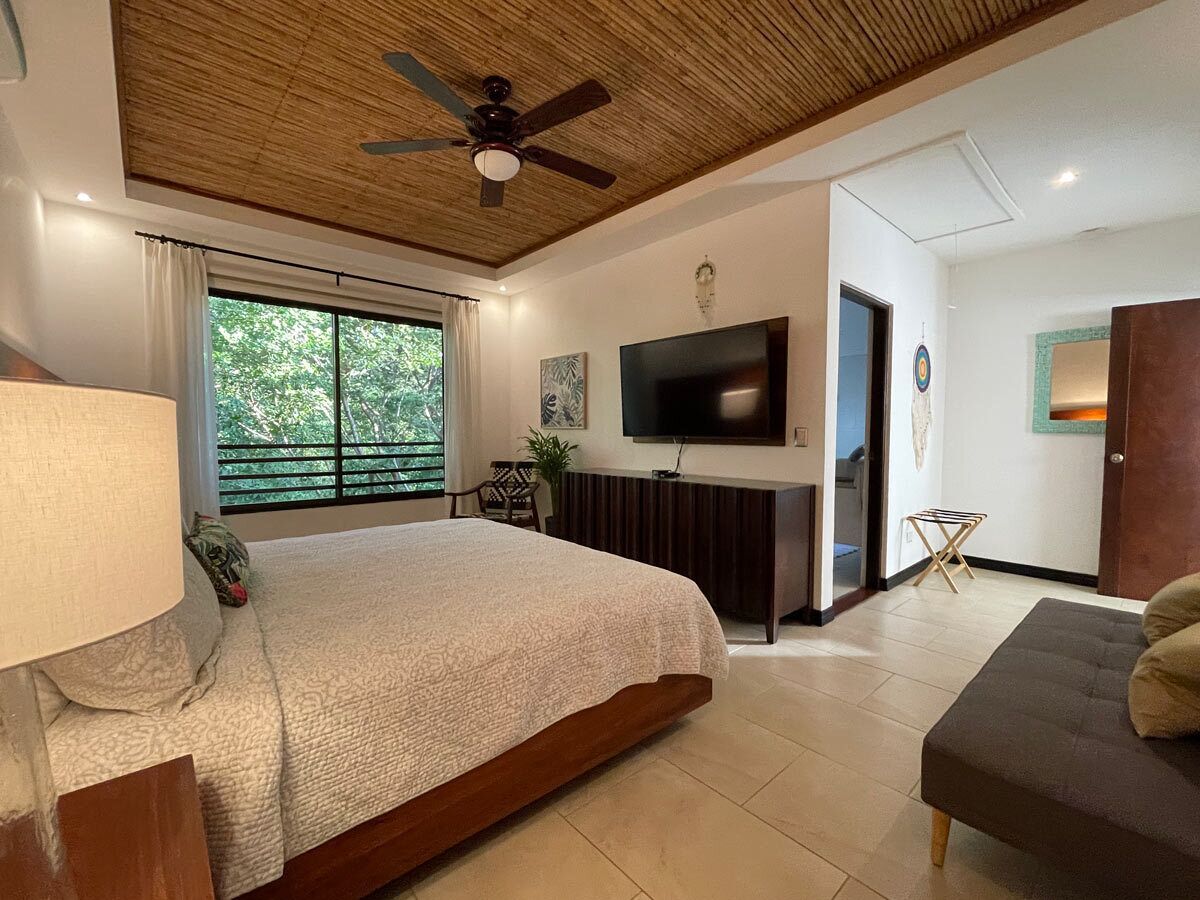 A bedroom with a large bed, a ceiling fan, a TV, and a view of greenery outside.