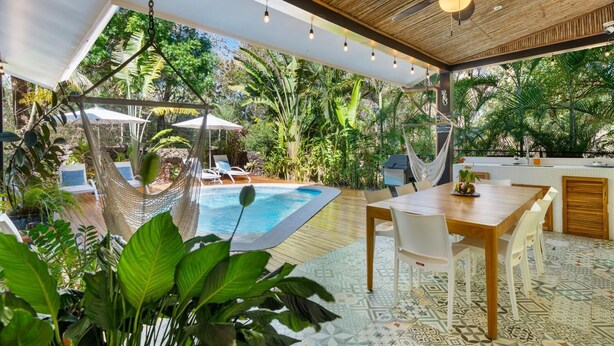 Un espacio exterior techado con piscina, sillas de descanso, una mesa de comedor y un ventilador de techo.