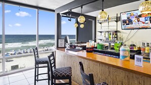 Un espace bar avec vue sur l’océan, des tabourets de bar, un comptoir avec un menu et un téléviseur diffusant les actualités.