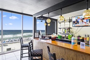 Een bar met uitzicht op de oceaan, barstoelen en een televisiescherm waarop nieuws wordt getoond.
