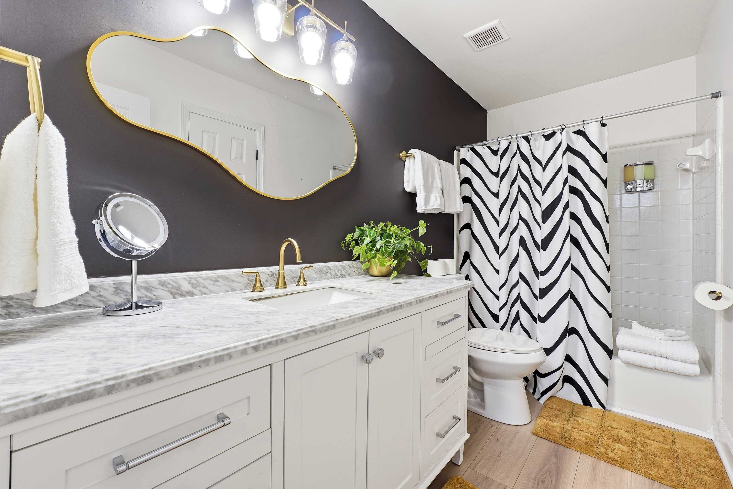 Une salle de bain moderne avec un comptoir en marbre, un miroir encadré d’or, des toilettes blanches et un rideau de douche à rayures noires et blanches.