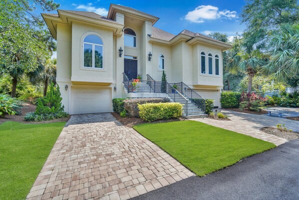 A two-story house with a brick driveway, a front lawn, and a staircase leading to a balcony.