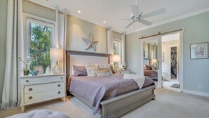 A bedroom with a large bed, a nightstand, a dresser, a ceiling fan, and a view of the outside.