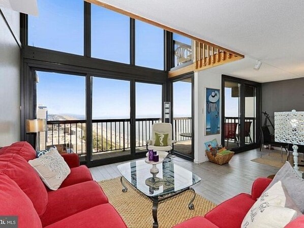 A spacious living area with a large glass table, red sofas, and a balcony with a view.