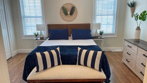 Ein Schlafzimmer mit einem Bett, zwei Nachttischen, einer Bank, einer Kommode und zwei Fenstern mit Jalousien.