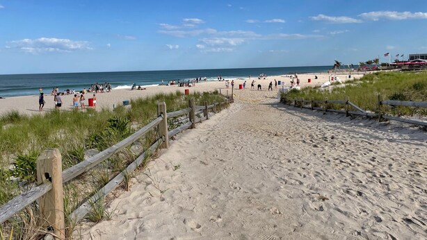 Ein Strand mit einer Holzzahlung, die zum Ufer führt, Menschen gehen spazieren und Strandstühle im Hintergrund.