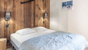 Een slaapkamer met een houten hoofdbord, twee wandlampen, een bed met wit beddengoed en een schilderij van een hert in een besneeuwd bos.