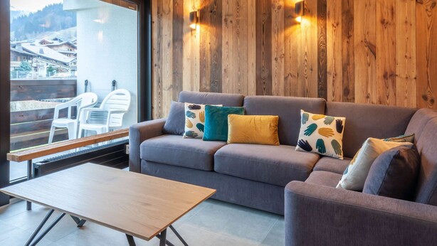 Een moderne woonkamer met een grijze bank, houten wanden en een balkon met witte stoelen.
