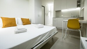 Quarto de hotel moderno com cama, escrivaninha e cadeira. Possui área de cozinha com armários e pia.