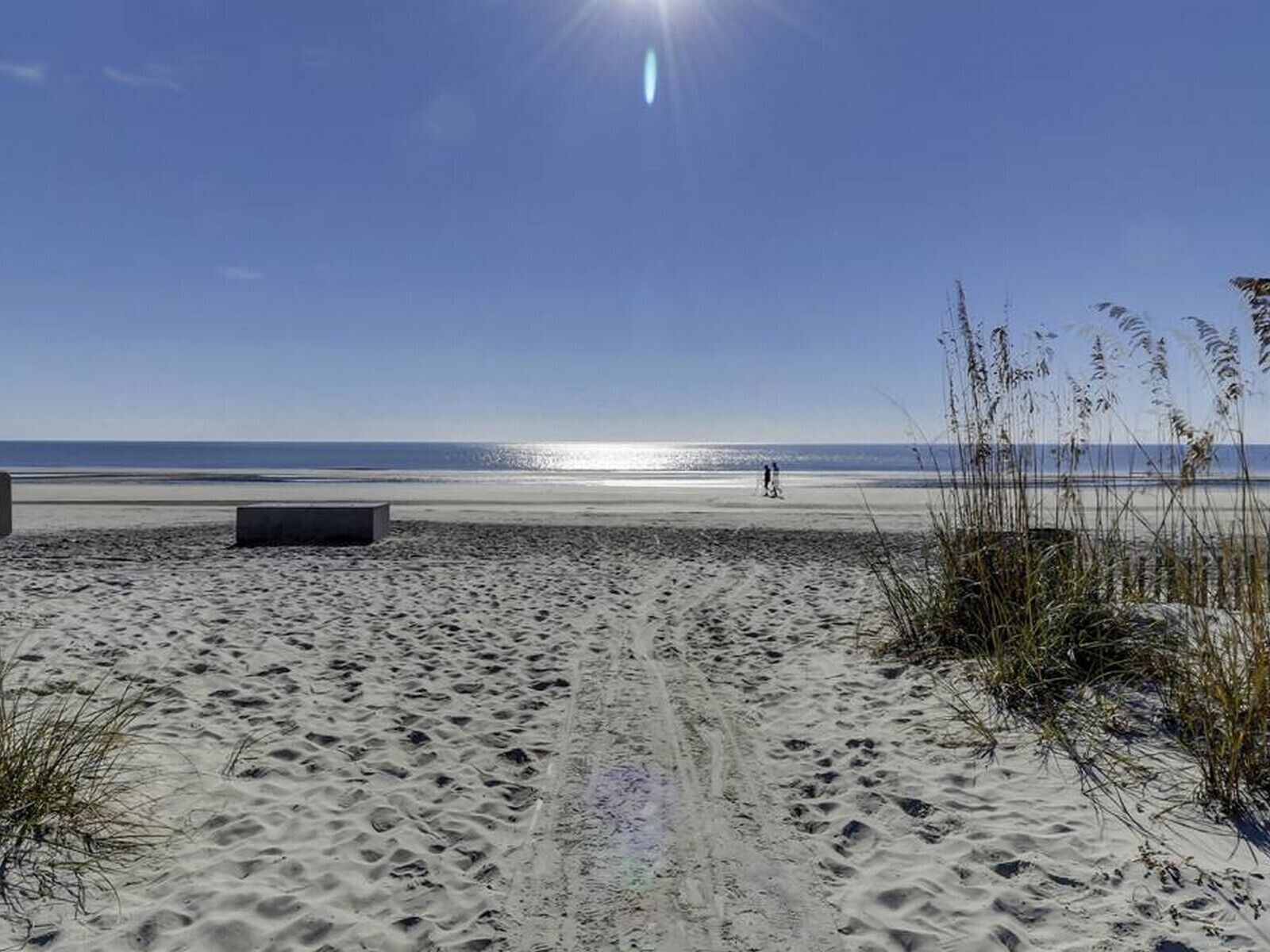 A sandy beach with footprints leading to the water, a clear sky, and a person walking in the distance.