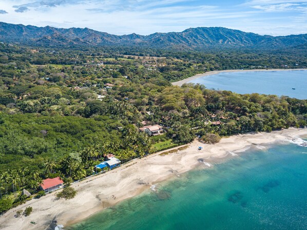 Vue aérienne d’une zone côtière avec une plage, une végétation luxuriante et un paysage montagneux en arrière-plan.