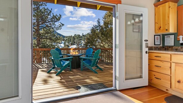 Une terrasse avec coin salon extérieur et foyer, une porte-fenêtre coulissante donnant sur un espace cuisine, et une vue sur les arbres et les montagnes.