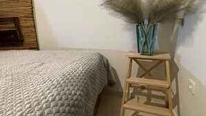 Un lit recouvert d’une couverture à motifs, un petit tabouret en bois surmonté d’un vase contenant des plumes d’herbe des pampas, et un mur avec un tableau de distribution électrique.