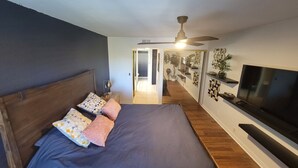 Ein Schlafzimmer mit Bett, Deckenventilator, Fernseher und einem Flur, der zu einem Badezimmer führt.