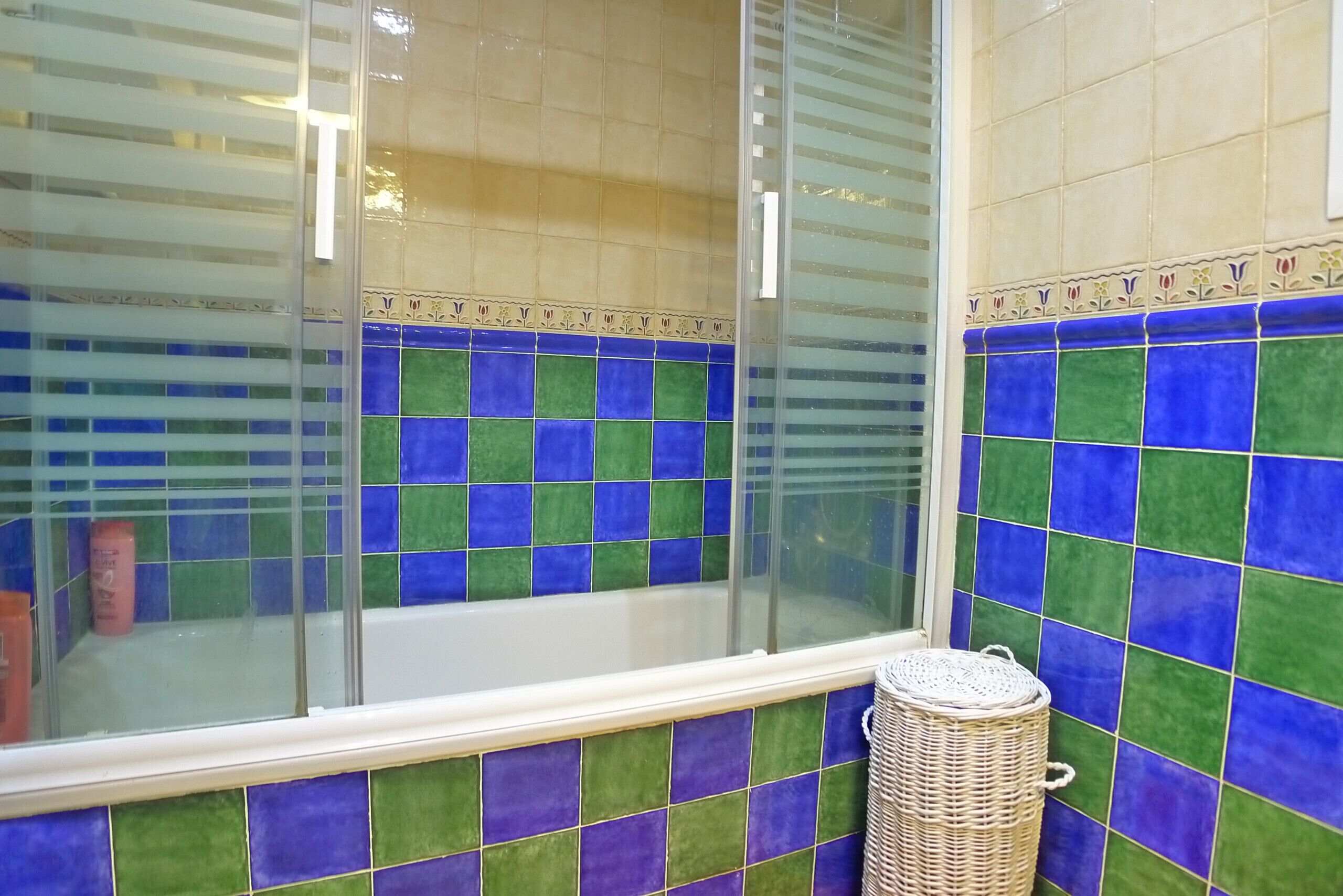 Un baño con bañera, paredes con azulejos azules y verdes, y una cesta de mimbre.