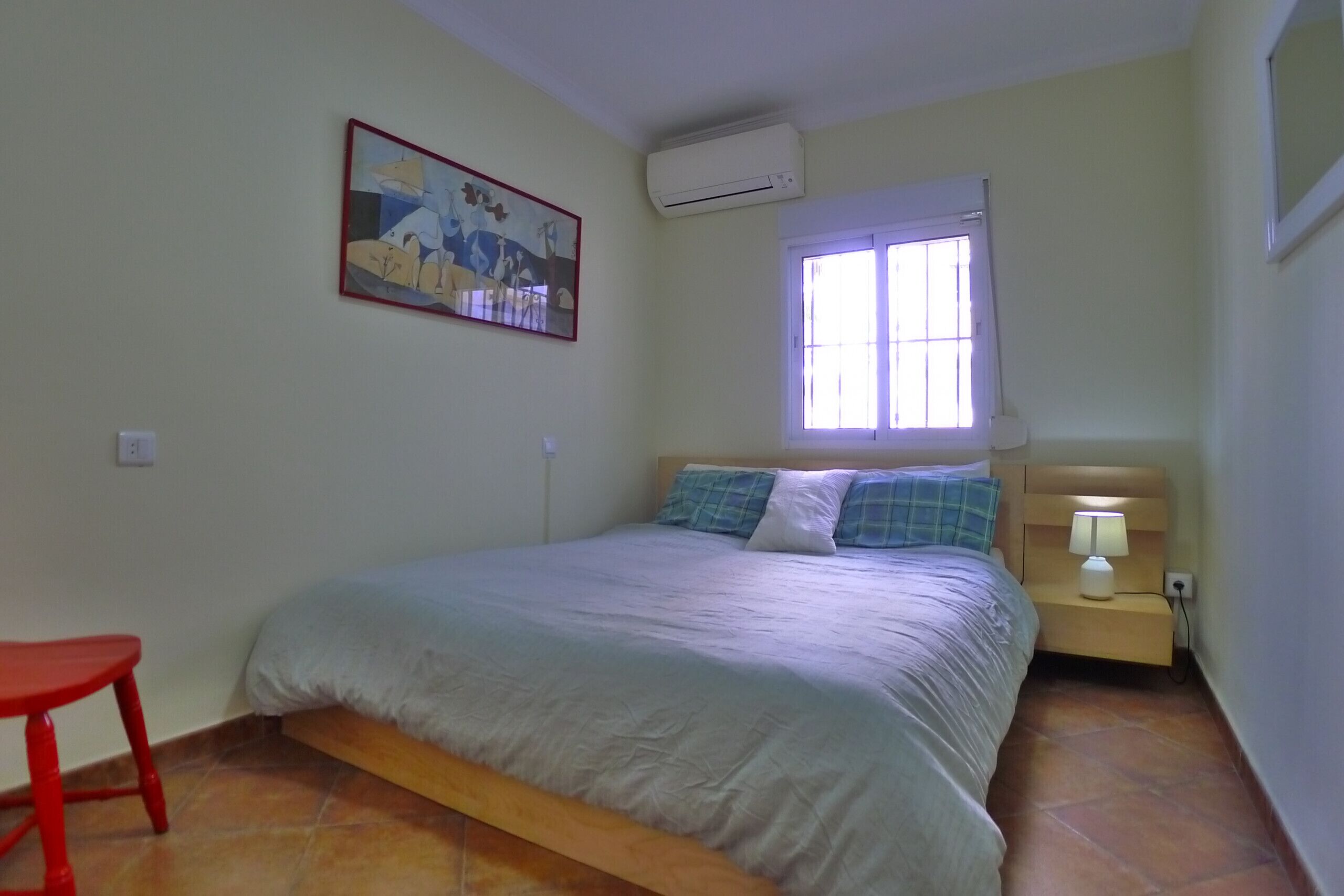 A bedroom with a bed, a nightstand with a lamp, a window with blinds, a red chair, and a wall-mounted air conditioner.