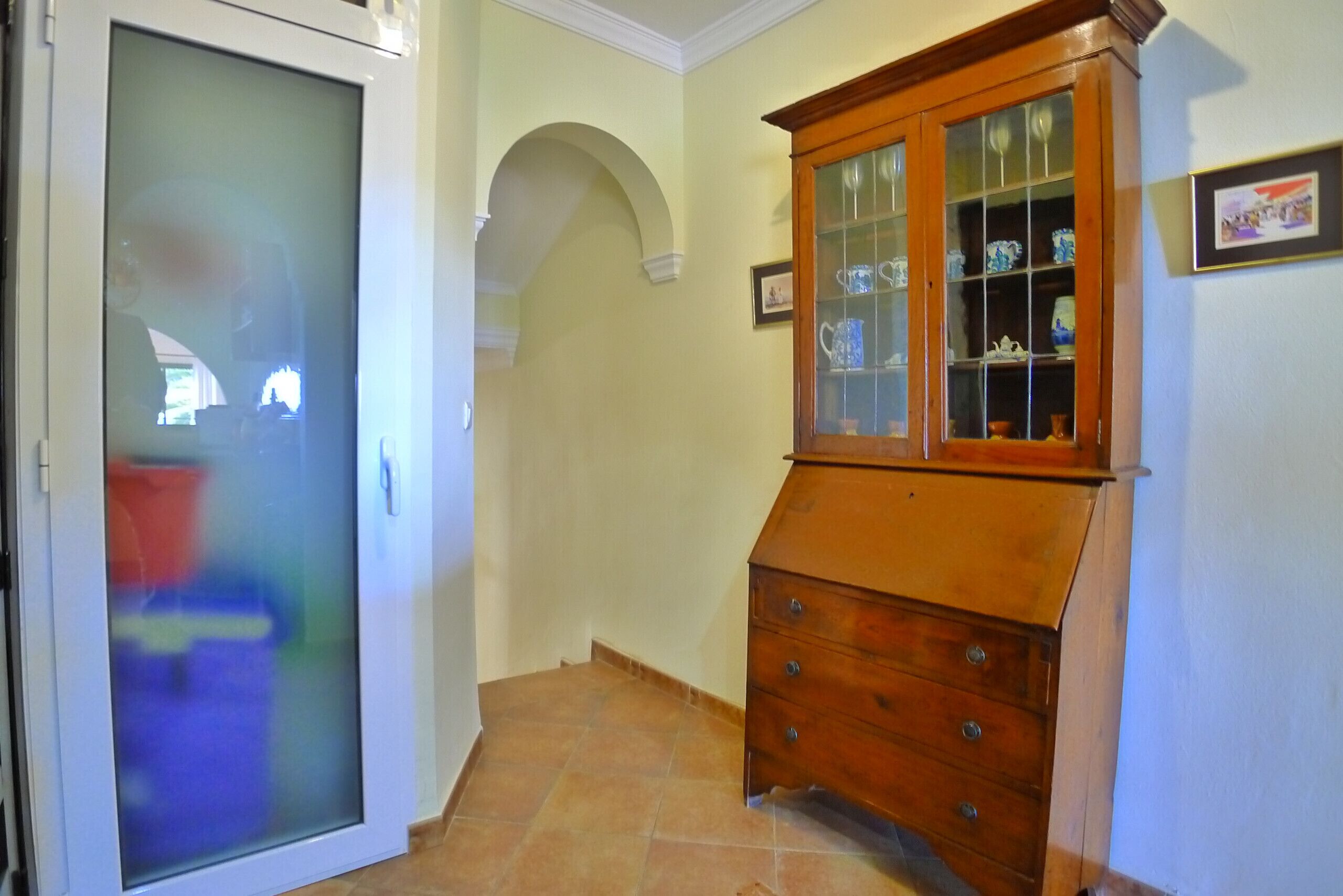 Un couloir avec une armoire en bois dotée de portes en verre, un sol carrelé et une porte blanche avec un panneau en verre dépoli.