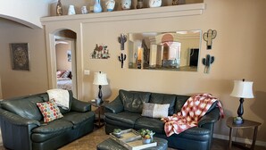 Ein Wohnzimmer mit zwei Ledersofas, einem Couchtisch und einem kleinen Beistelltisch mit Lampe. An der Wand hängt ein Spiegel, und ein Regal zeigt dekorative Gegenstände.