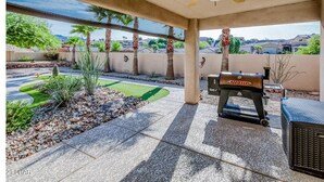 Eine überdachte Terrasse mit Grill, Deckenventilator und Blick auf einen angelegten Garten mit Palmen und Rasen.
