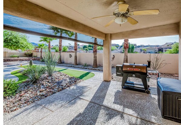 Un patio techado con una parrilla, un ventilador de techo y vistas a un jardín ajardinado con palmeras y un césped verde.
