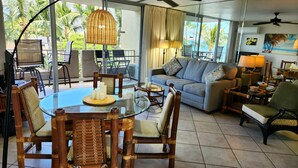 A spacious living area with a glass dining table, comfortable seating, and a view of palm trees. - Summer Special Pricing!  Oceanfront · Private Lanai · Heated Pool · Sunsets (Kihei)