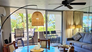 A spacious room with a round glass table surrounded by wooden chairs, a ceiling fan, and a large window offering a view of palm trees. - Summer Special Pricing!  Oceanfront · Private Lanai · Heated Pool · Sunsets (Kihei)