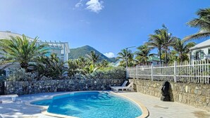 Una piscina con pared de piedra y sillas de descanso, rodeada de palmeras y una montaña al fondo.