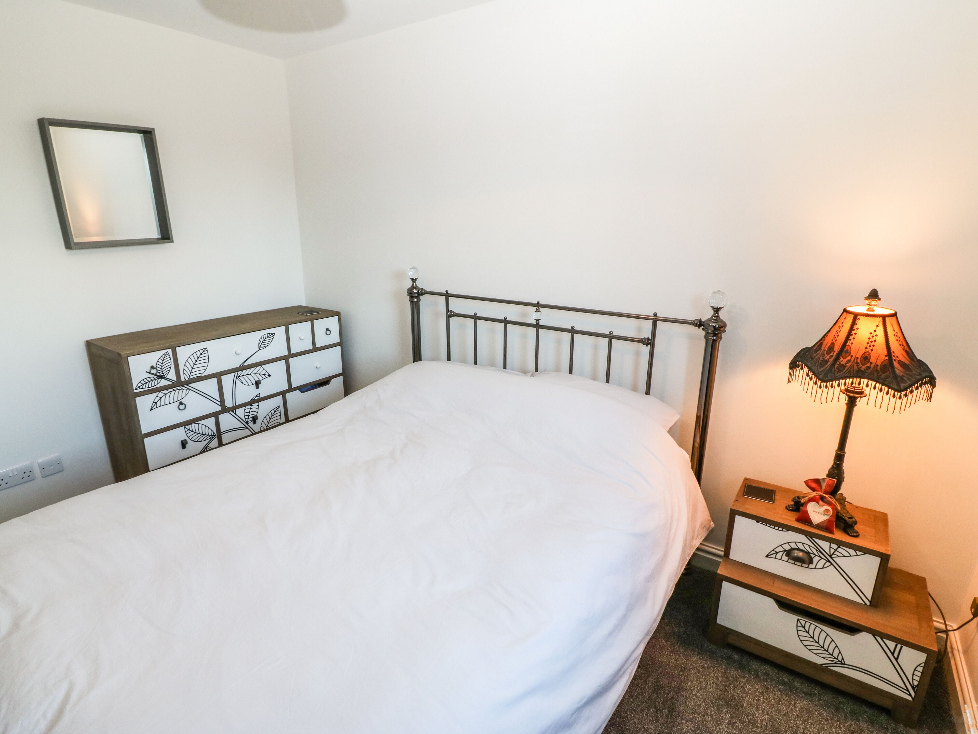 A neatly made bed with a metal frame, a bedside table with drawers, a lamp, and a wall-mounted picture frame.