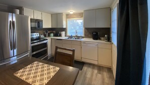 A modern kitchen with stainless steel appliances, a dining table with a geometric placemat, and a window with blinds. - Downtown Hub - Dog Friendly (Redmond)