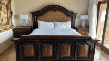 A large, ornate wooden bed with a headboard, flanked by nightstands with lamps, in a room with a tiled floor and a large window.