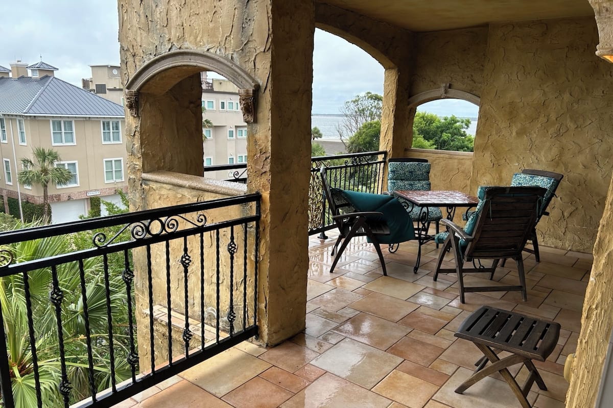 A balcony with a view of the sea, featuring a table and chairs, and a small wooden stool.