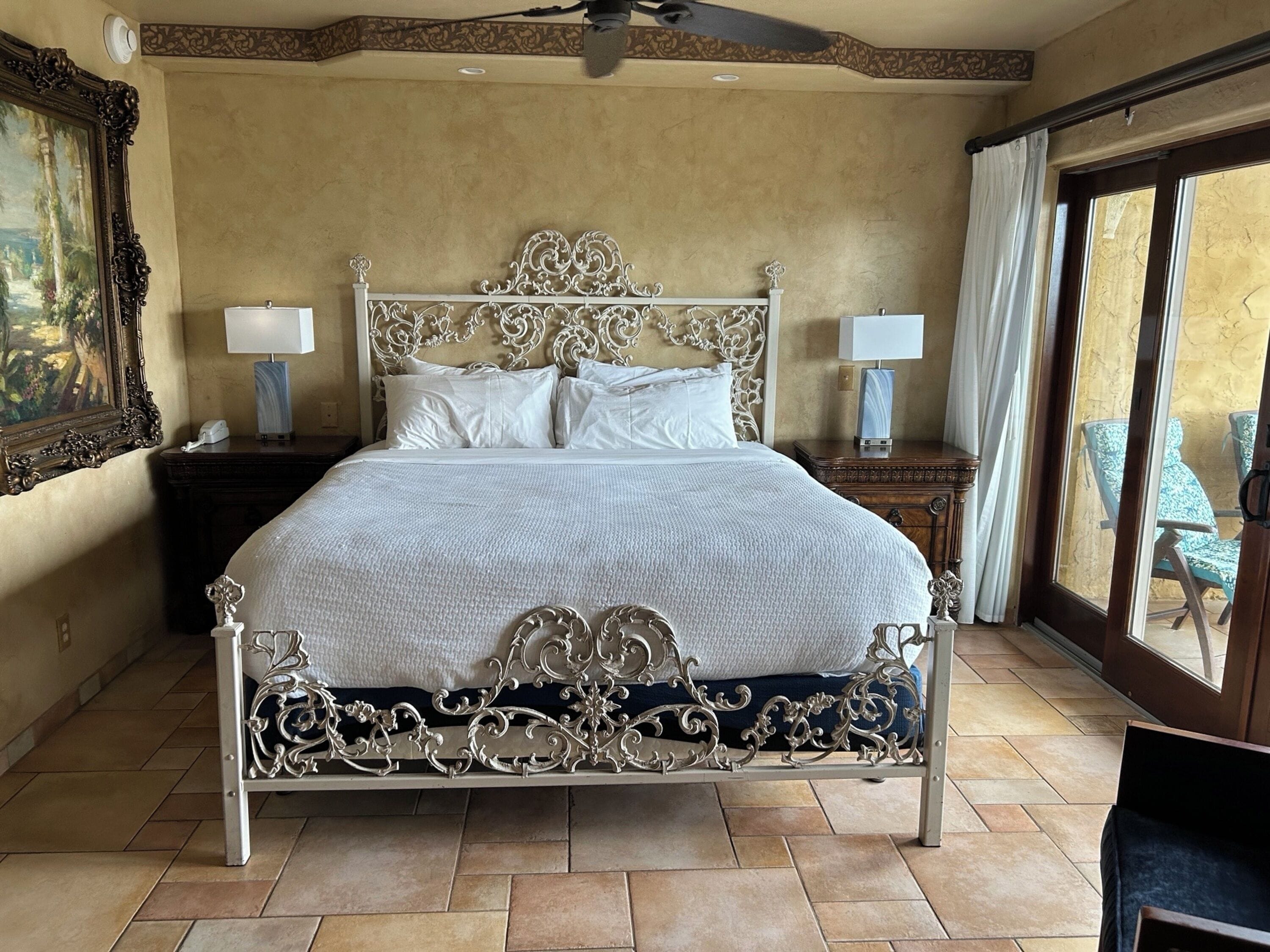 A spacious bedroom with a large bed, ornate headboard, two bedside tables with lamps, a ceiling fan, and a view of outdoor seating through a large window.