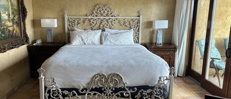 A spacious bedroom with a large bed, ornate headboard, two bedside tables with lamps, a ceiling fan, and a view of outdoor seating through a large window.