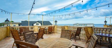 An outdoor patio with wooden furniture and string lights, overlooking a coastal area.