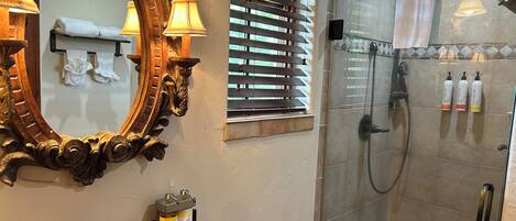 Un baño con un espejo grande y ornamentado, un lavabo con encimera negra y una zona de ducha con paredes de azulejos y puerta de cristal.