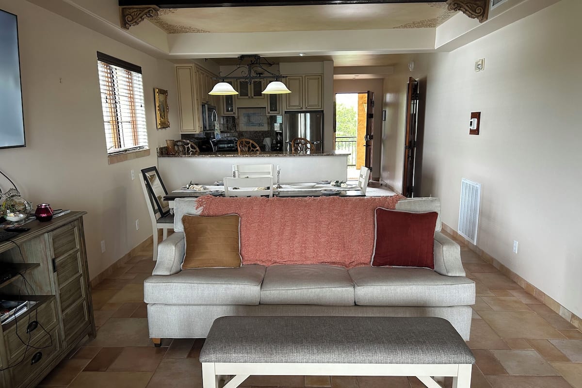 A spacious living room with a grey sofa, a white bench, and a kitchen in the background.